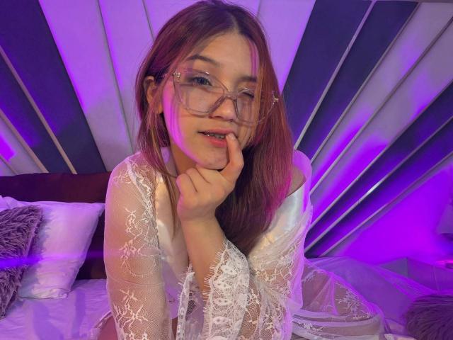 ElodieGardner with glasses, pink hair, and white lace robe, biting her finger, with purple lights. Her tits are small and she's looking cute and sexy.