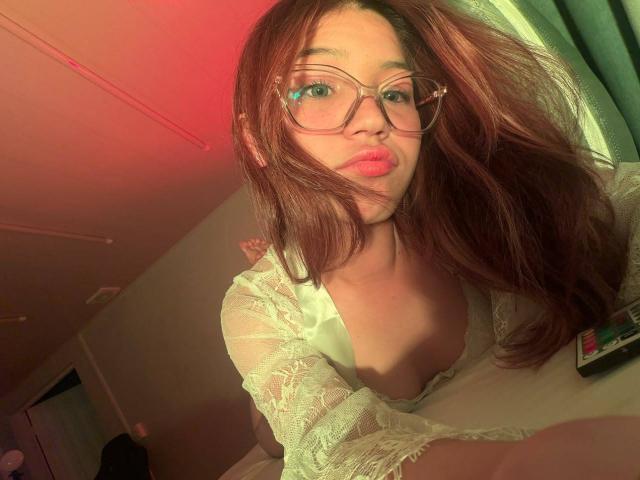ElodieGardner with red-tinted light, wearing glasses and a white lace top, pouting her pink lips, brown wavy hair, and showing her small tits in a selfie.