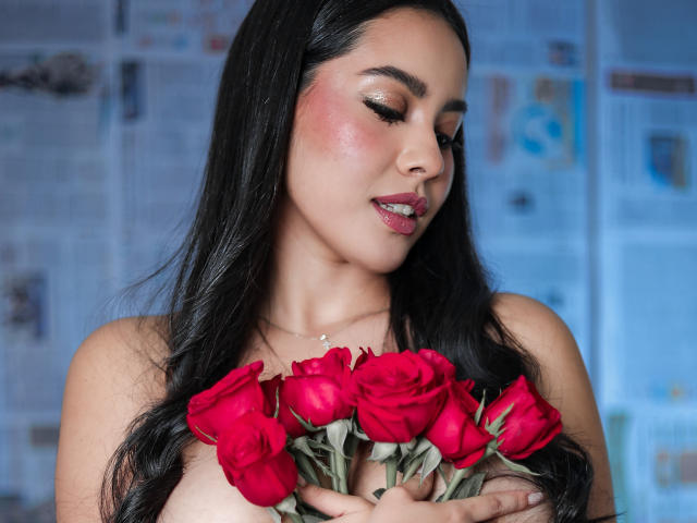PaolaYKatty met lang zwart haar, die rode rozen vasthoudt, haar borsten bedekt, kijkt naar beneden met een lichte glimlach, heeft roze lippenstift en glanzende oogschaduw. De achtergrond is wazig..