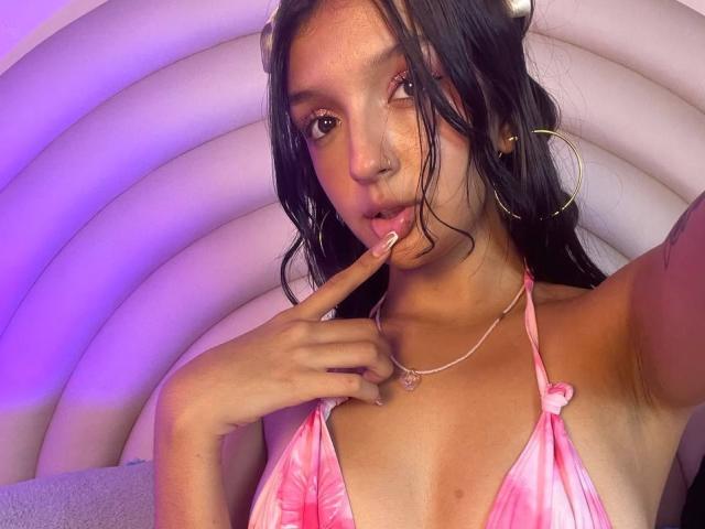 PaulineRey with long black hair, wearing a pink tie-dye bikini top, has a finger to her pink-tinged lips, large gold hoop earrings, and a heart necklace.