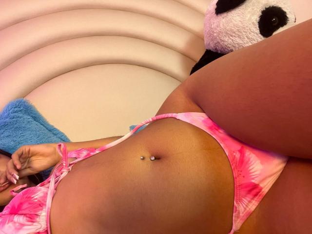 PaulineRey's tan skin, pink tie-dye bikini bottom, and belly button piercing are visible. Her ass is partially shown, with a blue and white stuffed panda in the background.
