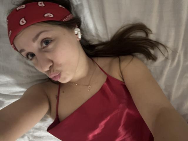 TrissyTerry with a red bandana, wearing a red satin camisole, pouting her lips, lying on white bed sheets, brown hair, blue eyes, white earbuds, necklace with small heart.