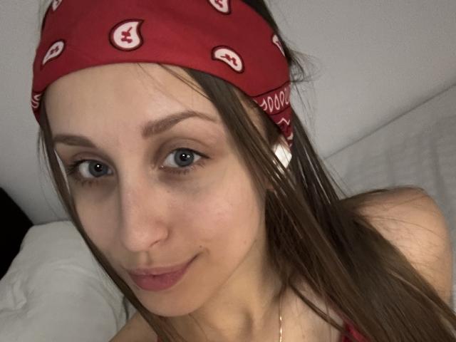 TrissyTerry with a red bandana, blue eyes, and brown hair, wearing a red top, has a cute smile and small nose, with a gold necklace visible.