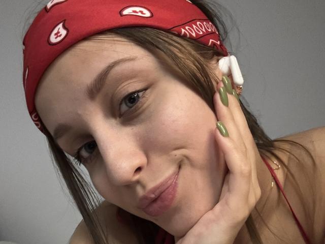 TrissyTerry with green nails, red bandana, and white earbuds, pouting slightly, resting her chin on her hand, showing a cute, confident expression. Her tits are small.