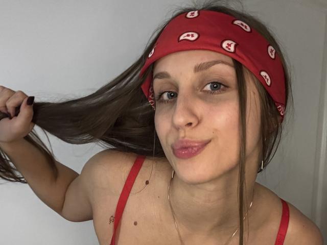 TrissyTerry with a red bandana, holding her brown hair, pink lips, blue eyes, and a small mole on her chest, wearing a red top, and a gold necklace.