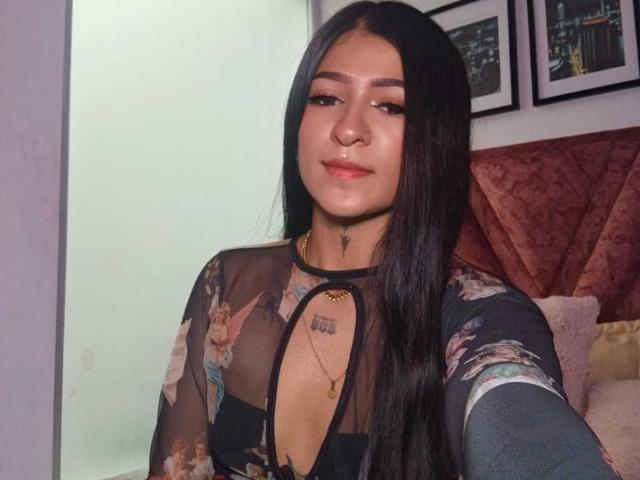 LuciadelaVega with long black hair, wearing a sheer floral top, showing her tits through the fabric, slight smile, gold necklace, tattoos on her chest, soft lighting.