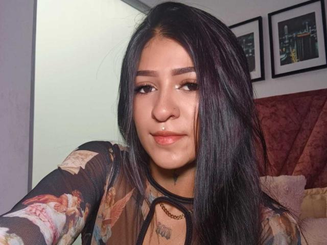 LuciadelaVega with long black hair, light brown skin, and a nose ring, wearing a sheer floral top, smiling slightly, looking to the side, in a bedroom.
