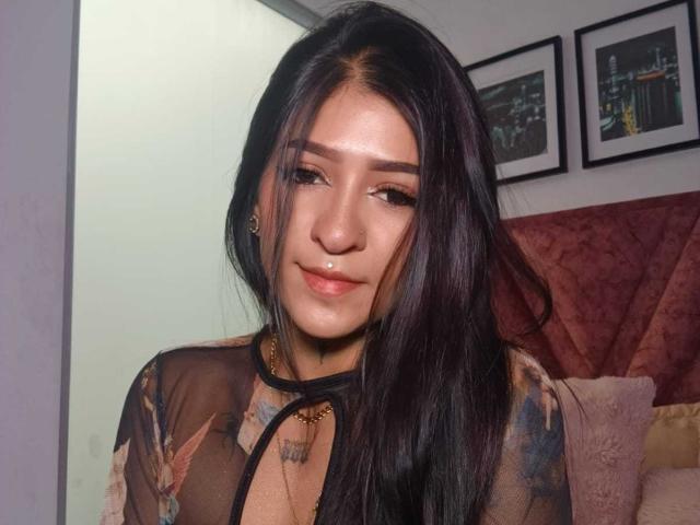 LuciadelaVega with long black hair, sheer top showing her small tits, light brown skin, subtle makeup, and a cute smile, standing in a bedroom.