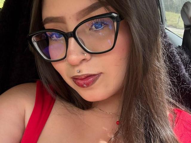 Madeleyn Rouse, com olhos azuis, óculos pretos, piercings no nariz e batom vermelho, vestindo uma blusa vermelha, com cabelo castanho e um colar, dentro de um carro. Seios parcialmente visíveis..