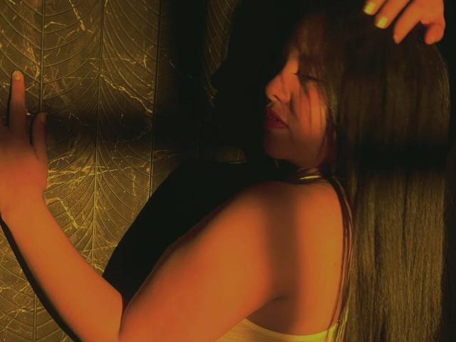 LilaVelvet with long brown hair, wearing a white tank top, leaning against a dark, textured wall with her right hand raised, her face partially shadowed, and her lips slightly parted.