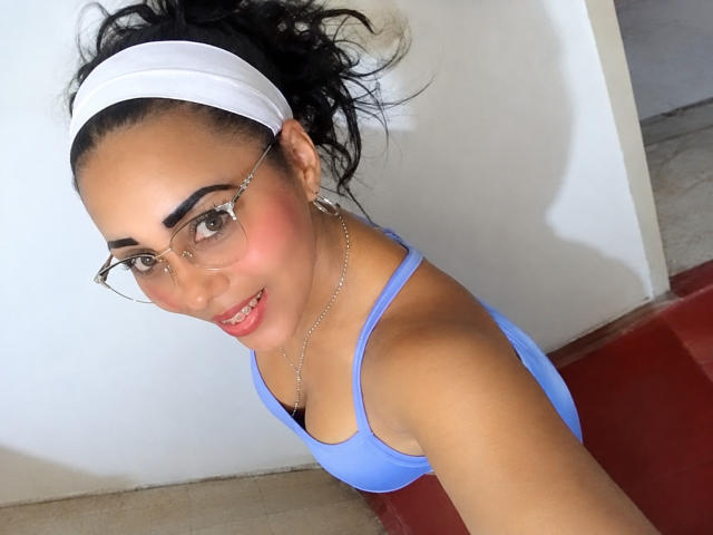 AvaAnyaa with dark curly hair in a white headband, wearing glasses, blue top, showing small tits, pink blush, smiling, necklace, red floor, beige wall.