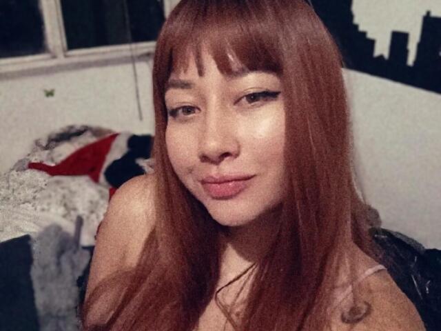 Genesiss with long red hair, bangs, and eyeliner, pouting lips, showing a cute smile, looking at the camera, in a bedroom with a messy bed.