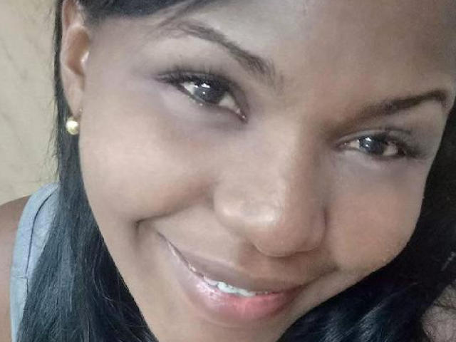 NayaDelaight's face, smiling with dark brown eyes, glossy black hair, small pearl earrings, and light brown skin, wearing a gray top, showing white teeth, slightly shiny lips.