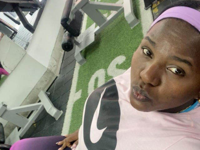 NayaDelaight with dark skin, pouty lips, pink headband, purple sports bra, black and pink Nike shirt, sitting in gym, looking up with a slightly confused expression.