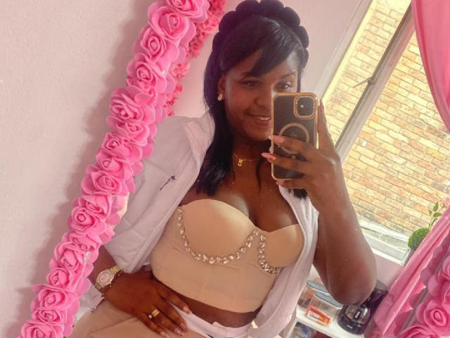 NayaDelaight with dark skin, black hair, wearing a strapless beige top with sparkly chains, white open shirt, pink roses mirror, holding phone, smiling, showing her tits.