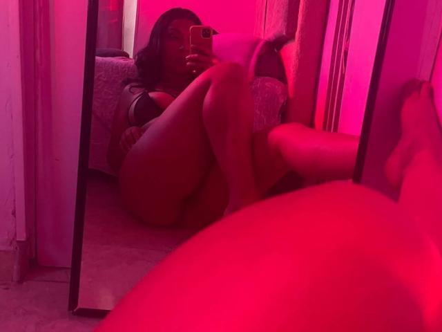 Sabrinags is taking a mirror selfie, showing her tits and ass, with red lighting, holding a phone in her right hand, sitting with legs up, and a bed in background.