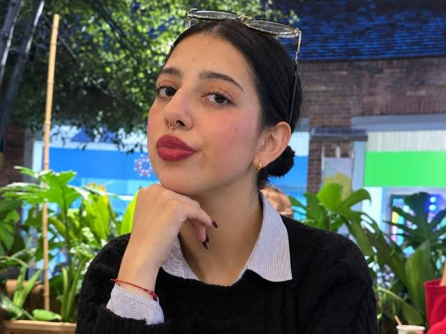 Hannah Cruz mit schwarzen Haaren, Nasenring und rotem Lippenstift, trägt einen schwarzen Pullover mit weißem Kragen, lehnt ihr Kinn in die Hand, trägt eine Sonnenbrille auf dem Kopf und ein rotes Armband..
