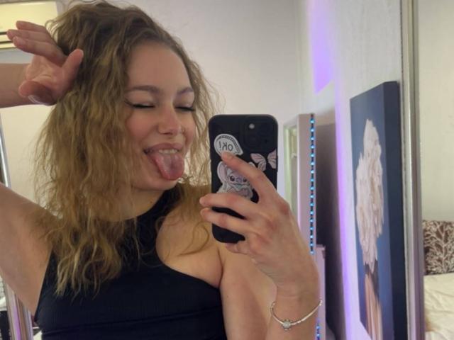 LeilaBie with wavy blonde hair, black sleeveless top, sticking out tongue, holding phone with "Grumpy Cat" sticker, mirror selfie, right arm raised. Boobs visible in reflection.