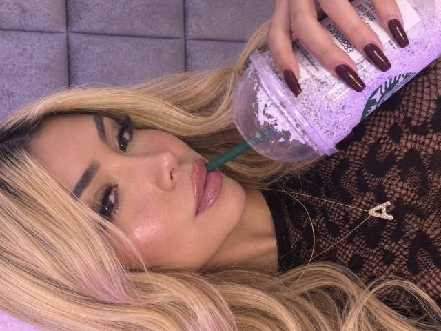 ChristieGarci with blonde hair, wearing lace top, sipping from a purple drink with a green straw, sporting dark red nails, and a gold cross necklace.