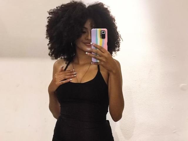 SnackTime with big, curly black hair, wearing a black tank top, holding a pink and yellow phone, smiling, showing off her small tits, standing in front of a white wall.