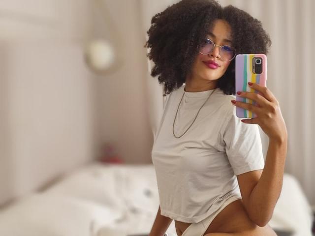 SnackTime, con cabello negro rizado, vestida con una camiseta blanca y ropa interior blanca, sosteniendo un teléfono con una funda de colores, tiene lápiz labial rosa y gafas, mirando a la cámara..
