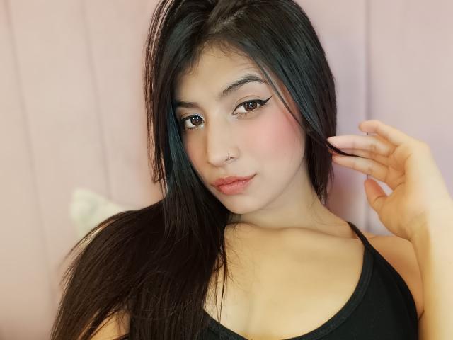 NaiaSanders with long black hair, nose piercing, and winged eyeliner, wearing a black tank top, has pink blushed cheeks, and a slight smile, touching her hair.