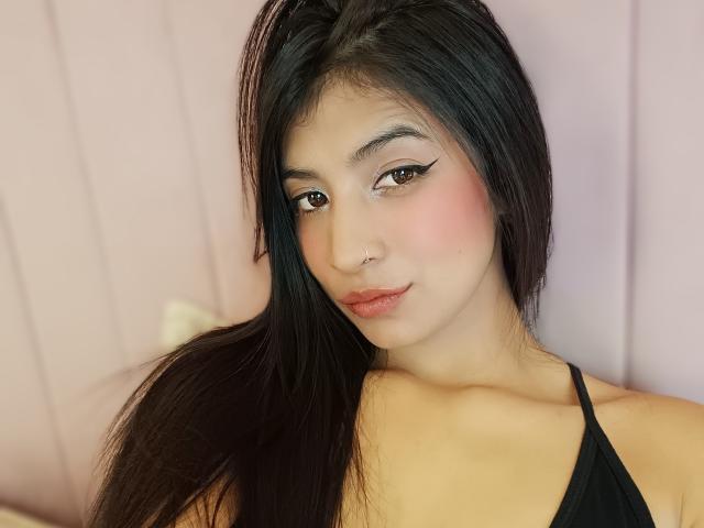 NaiaSanders has long black hair, brown eyes, pink blush, and a nose ring, wearing a black halter top, with a slight smirk and subtle makeup.