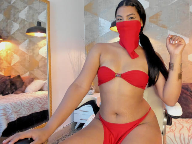 Lalila: Una ragazza moro con un bikini rosso, una maschera abbinata e una coda di cavallo nera, che mostra i suoi seni e i suoi glutei, seduta su una sedia in una camera da letto, con una mano vicino al viso..
