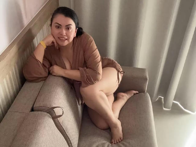 CharlotteDevereaux, curvy woman with dark hair, brown eyes, and light skin, lounges on a gray couch, wearing a brown, semi-sheer dress, showing her tits and ass.
