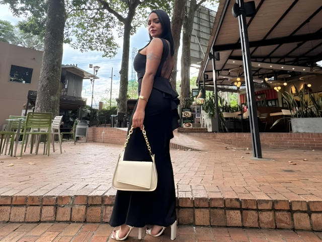 JessicaFiore with long black hair, wearing a black dress, holding a cream handbag, standing on a brick patio, looking back, showing her ass, wearing white heels.