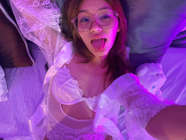 ElodieGardner with glasses, tongue out, wearing white lace lingerie and robe, pink and purple lighting, showing small tits and a cute face, lying on bed with plush toy.