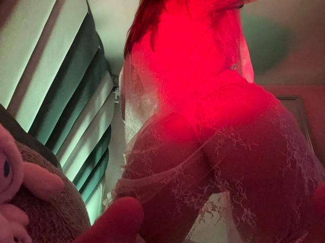 ElodieGardner's ass in lacy white lingerie, illuminated by red light, with her back to the camera, her legs slightly apart, and a pink stuffed animal on the left.