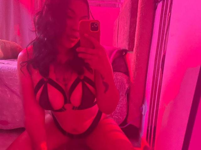 Sabrinags with wavy dark hair, wearing a black strappy bra, sitting on the floor, holding a phone, red-pink lighting, visible tattoo on her arm, mirror selfie.