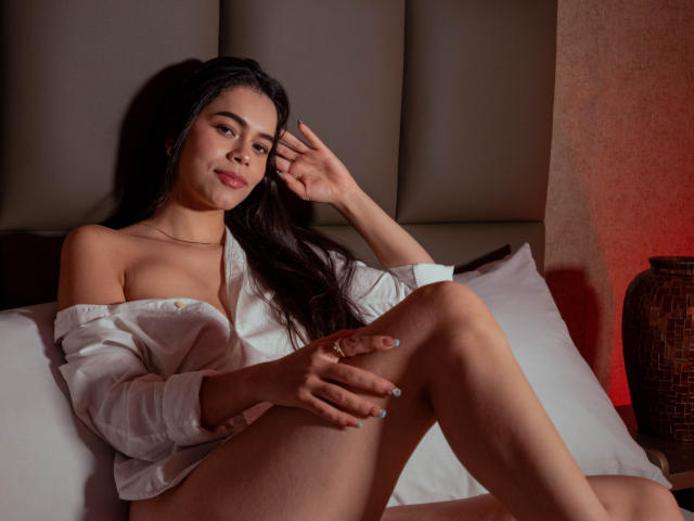 AlanaBlake with long dark hair, white off-shoulder shirt, and no pants, sitting on a bed, legs up, hand touching her head, looking at the camera, smiling.