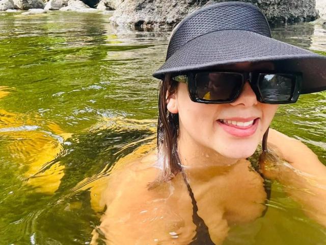 CloeEvanss, con i capelli castani bagnati, indossa occhiali da sole neri e un berretto nero, sorridendo in acqua verde, mostrando i seni appena sopra la superficie. Una roccia sullo sfondo..