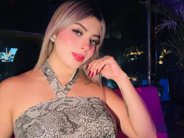 CloeEvanss, con cabello rubio, lápiz labial rojo y un vestido de tirantes con estampado de serpiente, mostrando sus senos y esmalte de uñas rojo, con la mano cerca de su rostro. Fondo de discoteca..