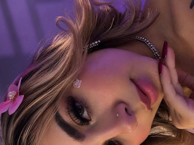 CloeEvanss's face with blonde hair, pink flower, diamond headband, red nails, nose piercing, and makeup. She's lying down with purple lighting, looking at the camera.
