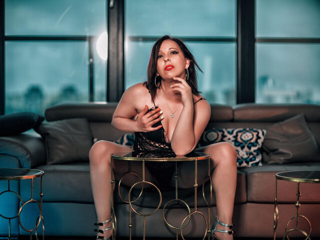 LauraSin with medium tits, sitting on a gold circular table, wearing a black lace dress, legs spread, hand on chest, red lipstick, city lights through windows in the background.