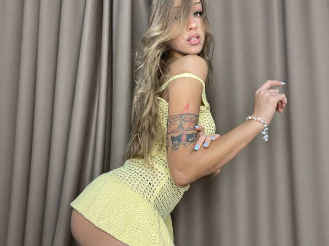 Helena-hot with long blonde hair, yellow crochet dress, showing her ass, tattoo on right arm, and a bracelet, standing against beige curtains.
