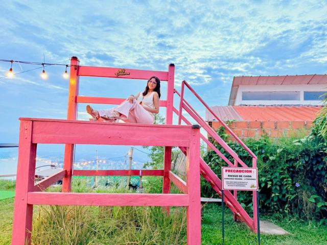 HanaJoud lounges on a bright pink wooden chair, wearing a white dress, with her legs up, showing her ass, against a cloudy blue sky, surrounded by greenery.