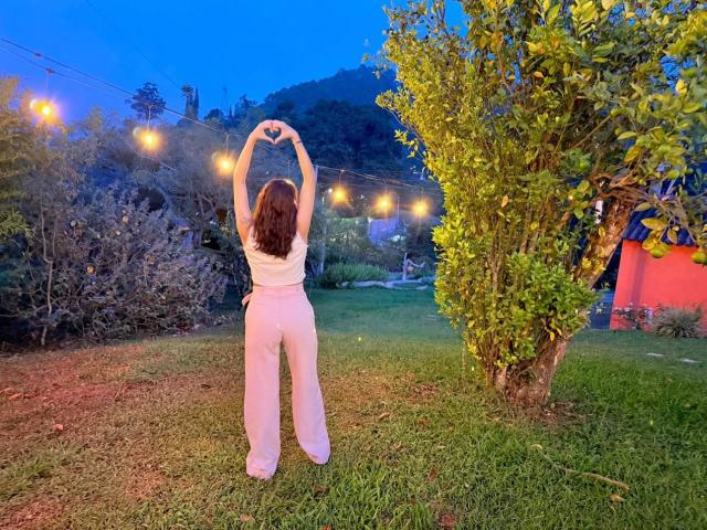HanaJoud stands with arms raised, back to camera, wearing a white top and pink pants, in a grassy yard with string lights, blue evening sky, and a tree on the right.