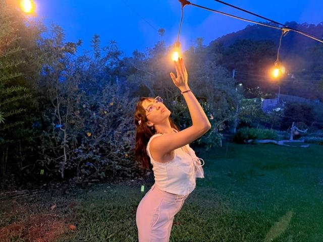 HanaJoud with glasses, white top, and white pants, stretching up to touch glowing string lights, blue evening sky, green grass, and bushes in the background.
