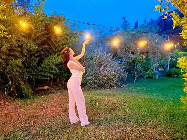 HanaJoud with her tits up, reaching for a glowing string light, wearing a white, sleeveless, high-waisted, flared pants outfit, in a grassy yard at dusk.