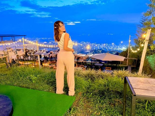 HanaJoud stands with her back to the camera, wearing white pants and a sleeveless top, looking over her shoulder, showing her ass, in a rooftop bar with string lights.