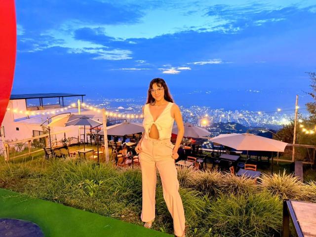 HanaJoud stands in a white, cut-out, sleeveless jumpsuit with high-waisted pants, hands in pockets, on a grassy hill, blue evening sky, and a city lights view behind her.