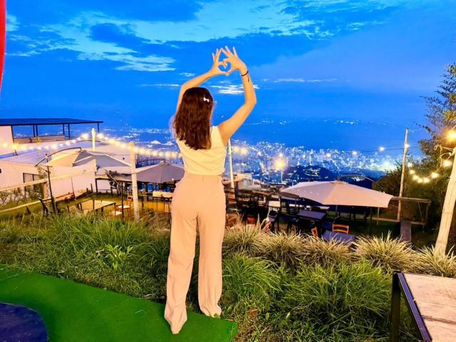 HanaJoud stands with arms raised, hands forming a circle, facing a city skyline at dusk. She's wearing a white sleeveless top and beige pants, with green grass below.