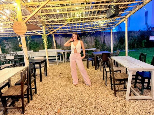 HanaJoud with long brown hair, wearing a white tied-up top and pink high-waisted pants, stands in a gravel-covered patio, talking on a phone, under a wooden pergola.