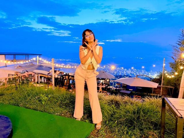 HanaJoud stands with hands near her face, wearing a white sleeveless top and beige flared pants, against a twilight sky. Her ass is visible under her top.