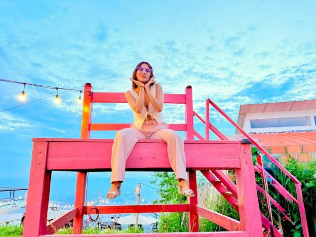 HanaJoud sits on a pink wooden ladder, wearing a beige top and pants, talking on a phone, with string lights above and a blue sky in the background.