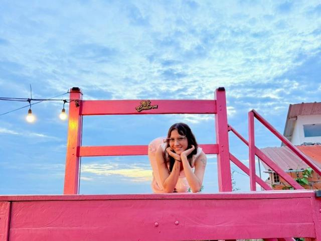 HanaJoud crouches on a pink wooden railing, smiling with hands on chin, wearing white, long black hair, blue sky with clouds, string lights, and a pink building in background.